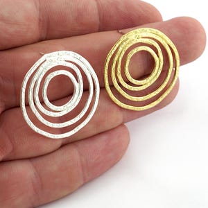 Op de afbeelding: Twee ronde oorknopjes, één in zilverkleur en één in goudkleur. Elke oorbel heeft een reeks van drie concentrische ringen met een getextureerde, gehamerde oppervlakteafwerking, wat zorgt voor een gelaagd, opengewerkt ontwerp.