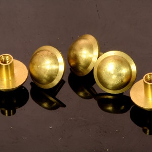 Puede incluir: Cinco sujetadores de metal de color dorado en forma de cúpula con un poste roscado. Los sujetadores tienen una base plana y una parte superior abovedada.