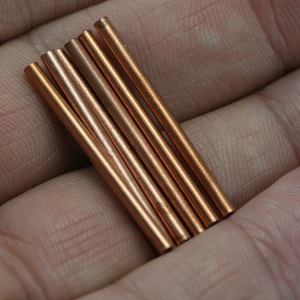 Könnte beinhalten: Sechs Kupferstäbe, in einem Stapel angeordnet, werden in einer Hand gehalten. Die Stäbe sind glatt und glänzend.