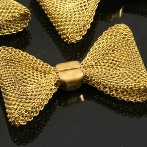 May include: A gold metal bow with a triangular shape. The bow is made of a chain-like material and has a small, round, brass-colored connector in the center.