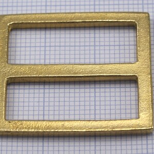 May include: A gold-colored metal buckle with two rectangular openings. The buckle has a smooth, slightly textured surface.