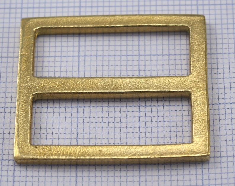 May include: A gold-colored metal buckle with two rectangular openings. The buckle has a smooth, slightly textured surface.