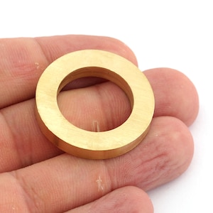 Könnte beinhalten: Ein massiver Messingring mit kreisförmiger Form. Der Ring hat eine flache Oberfläche und ein Loch in der Mitte. Der Ring wird in einer Hand vor weißem Hintergrund gehalten. Der Ring hat eine warme Goldfarbe.