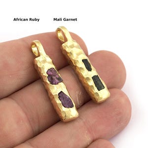 May include: Two gold-colored pendant necklaces with textured surfaces. One pendant features rough-cut African Ruby stones, and the other has Mali Garnet stones. Both pendants have a loop at the top for a chain.