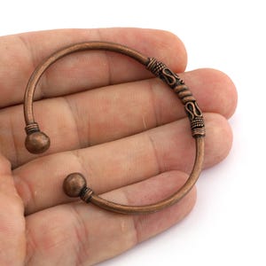 May include: Copper-colored cuff bracelet with decorative detailing. The open-ended bracelet features a rounded design with ball-shaped ends and an ornate central section. The bracelet is designed to be worn on the wrist.