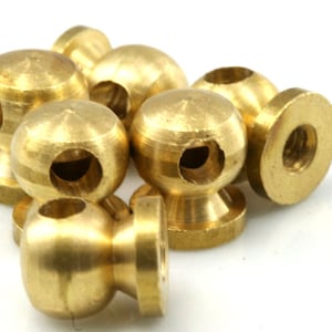 Puede incluir: Un conjunto de pequeños pomos esféricos de color latón, cada uno con un agujero perforado a través de la esfera. Los pomos tienen una base redondeada y probablemente se utilizan para muebles o fines decorativos. El hardware se muestra sobre un fondo blanco.