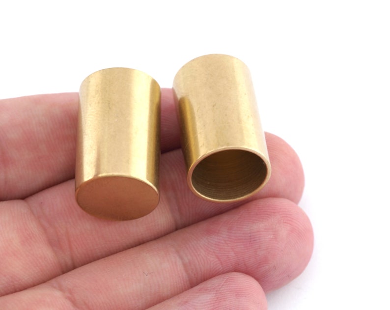 May include: Two cylindrical metal components with a smooth, matte gold-toned finish. One piece is positioned with its solid flat end facing upward, while the other is oriented to reveal its hollow, open interior and thin-walled construction.