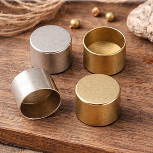 May include: Four brass cord end caps in antique silver and raw brass finish are displayed on a wooden surface. The caps are cylindrical, with two in silver and two in gold. The text "Antique Silver & Raw Brass Brass Cord End Caps" is at the top. The text "14x10mm | Nickel Free" is at the bottom.