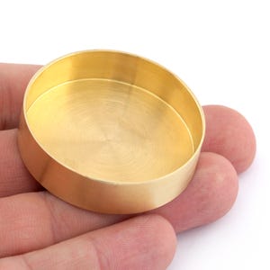 Puede incluir: Un pequeño recipiente redondo de latón con una superficie lisa y brillante. El recipiente está vacío y parece estar hecho de latón macizo.