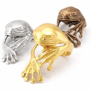 Op de afbeelding: Drie kikker-vormige ringen in zilver, goud en brons. De ringen hebben gedetailleerde kikkerontwerpen met gesloten ogen en uitgestrekte poten. De ringen zijn gemaakt van metaal en worden op een witte achtergrond getoond.