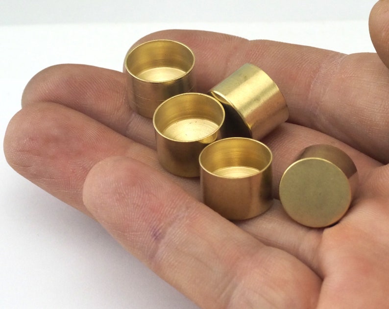 May include: A collection of small, cylindrical brass components. The objects are golden in color and have a smooth, reflective surface. The components are held in a hand, showcasing their size and shape. They appear to be small containers or decorative elements.