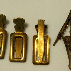 Può includere: Quattro fermagli per capelli in metallo dorato. Tre sono rettangolari con una piccola linguetta appuntita al centro. Uno è un fermaglio più lungo e curvo con un bordo dentellato.