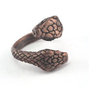 Könnte beinhalten: Ein kupferfarbener Ring mit einem Design aus einer zweiköpfigen Schlange. Die Schlangenköpfe sind in entgegengesetzte Richtungen gerichtet und haben eine strukturierte, schuppige Oberfläche.