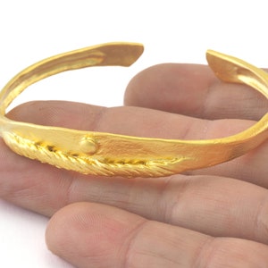 以下が含まれることがあります： ロープ状のデザインと小さな丸いアクセントが付いた金のバングルブレスレット。ブレスレットはオープンタイプで、ハンマード仕上げになっています。