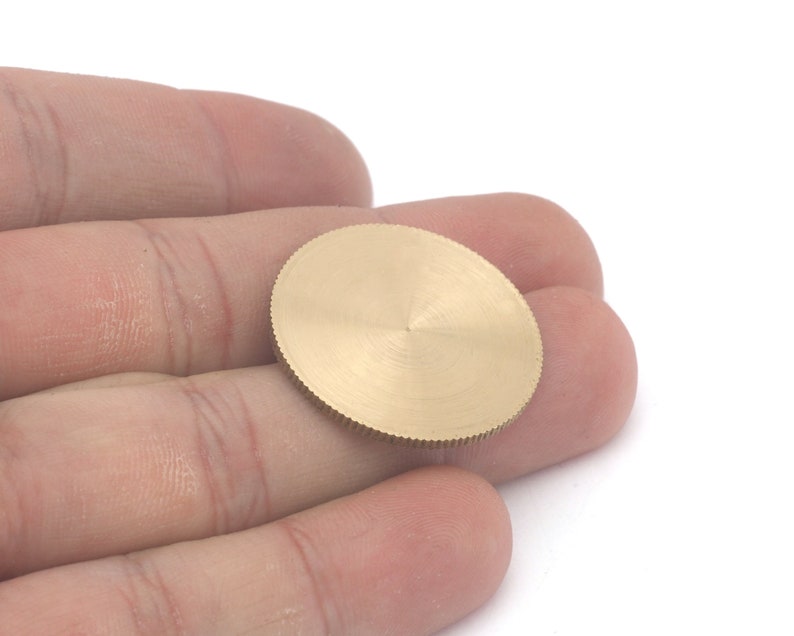 Op de afbeelding: Een klein, goudkleurig, cirkelvormig metalen object met een geribbelde rand en een getextureerd oppervlak. Het object wordt in de hand van een persoon gehouden tegen een witte achtergrond. Het lijkt een kleine schijf of component te zijn.