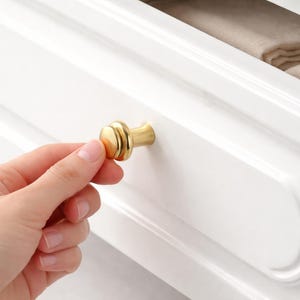 Puede incluir: Una taza de cerámica blanca con un asa negra y el texto "But First, Coffee" en un estilo manuscrito. La taza está sobre una superficie blanca. La taza mide 10 cm de alto.