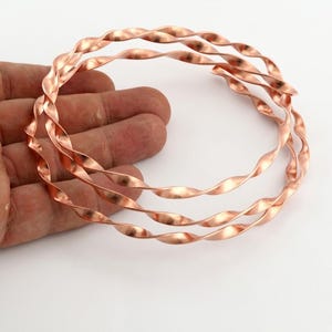 Op de afbeelding: Een set van drie cirkelvormige armbanden gemaakt van gepolijst koperdraad. Elke armband heeft een doorlopend gedraaid spiraalontwerp dat een golvende, glooiende textuur creëert langs de gehele omtrek van de metalen banden.