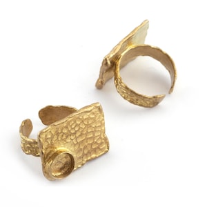 May include: Two gold-toned rings featuring a rectangular, hammered-texture face. Each ring has an open, adjustable band with a rough, organic finish and a circular bezel setting positioned on the corner of the rectangular plate.