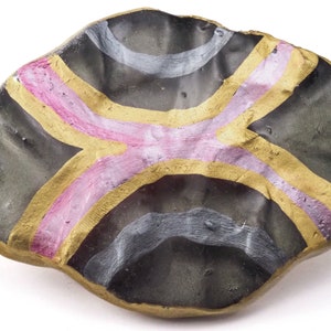 May include: A handcrafted, irregularly shaped decorative dish. The dish features a dark gray base with a gold-colored rim and accents. A pink and gold cross-like design is painted across the center, with additional gray and white curved lines.