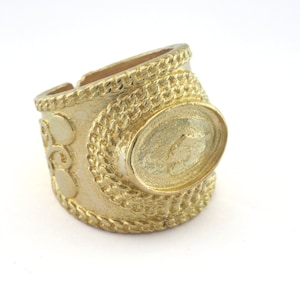Op de afbeelding: Een ring van goudkleurig metaal met een ovaalvormig ontwerp en ingewikkelde details. De ring heeft een getextureerde band met een touwachtig patroon en een verhoogd ovaal in het midden.