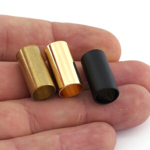 May include: Three metal tubes, one gold, one brass, and one black, are shown in a hand. The tubes are all the same size and shape.