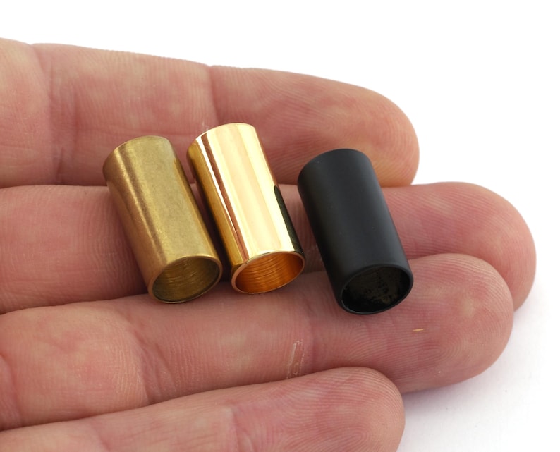 May include: Three metal tubes, one gold, one brass, and one black, are shown in a hand. The tubes are all the same size and shape.