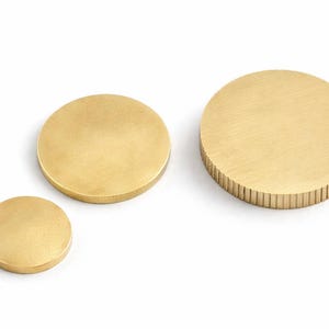 Op de afbeelding: Drie messing cirkelvormige objecten van verschillende afmetingen worden getoond tegen een witte achtergrond. De kleinste is een massieve schijf, de middelste is glad en de grootste heeft een geribbelde rand. Deze decoratieve items kunnen worden gebruikt als onderzetters of decoratieve elementen.