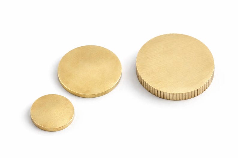 Op de afbeelding: Drie messing cirkelvormige objecten van verschillende afmetingen worden getoond tegen een witte achtergrond. De kleinste is een massieve schijf, de middelste is glad en de grootste heeft een geribbelde rand. Deze decoratieve items kunnen worden gebruikt als onderzetters of decoratieve elementen.