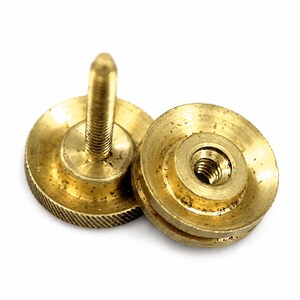 May include: Two circular brass fasteners with a metallic gold finish. One fastener features a central threaded post extending upward, while the other displays a central threaded hole. Both components have a textured, knurled edge for grip.