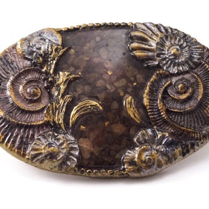May include: An oval-shaped, decorative object with a dark brown center filled with small, light brown stones and gold flecks. The edges are dark brown with gold and silver accents, featuring detailed, spiraling fossil designs.