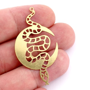 Puede incluir: Un amuleto de metal dorado con una serpiente enroscada alrededor de una luna creciente. La serpiente y la luna están recortadas en el metal, creando un diseño delicado e intrincado.