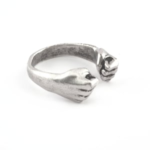 May include: A silver ring with two hands clasped together, forming a circle. The hands are detailed and realistic, with visible fingers and knuckles.