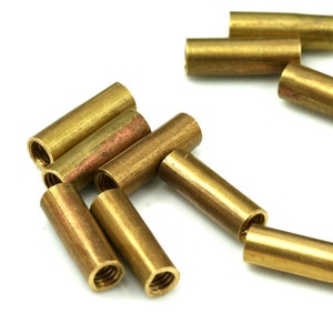 Puede incluir: Un grupo de insertos roscados de latón, también conocidos como separadores, están dispersos sobre una superficie blanca. Los insertos son cilíndricos y tienen un agujero roscado en un extremo.