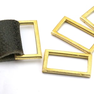 Puede incluir: Cuatro marcos rectangulares de metal de color dorado, algunos están apilados uno encima del otro. Un marco está unido a un trozo de cuero marrón.