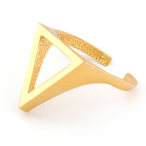 May include: A gold-colored ring with a geometric triangle design. The ring features a textured band and a smooth, solid triangle shape. The ring is set against a white background, highlighting its modern and minimalist style.