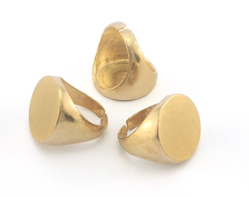 May include: Three gold-colored adjustable signet rings with a matte finish. Each ring features a round, flat face and a simple band. The rings are displayed on a white background, showcasing their design.