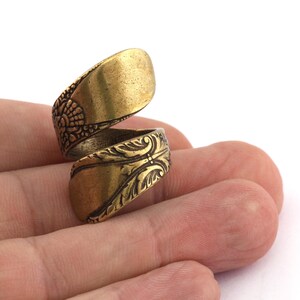 May include: A gold-toned ring made from a twisted spoon with an ornate design. The ring is on a finger.
