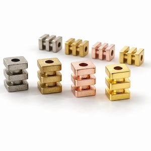May include: An assortment of metal beads in silver, gold, and copper tones. The beads have a square shape with a hole in the center and are stacked in a three-tiered design. Some beads have a three-bar design.