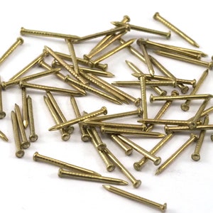 May include: A pile of gold-colored tacks or upholstery nails. The tacks are scattered on a white surface.
