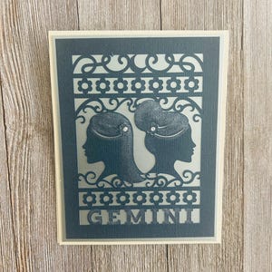 Może przedstawiać: Karta w ciemnoniebieskim kolorze z wyciętym wzorem dwóch postaci kobiecych z napisem "GEMINI" na dole. Karta ma białą ramkę.