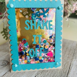 May include: A teal blue card with a scalloped border and white dots. Inside the card is a gold background with colorful sequins and the words "SHAKE IT OFF" in blue.