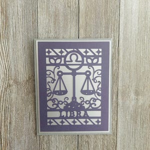 Może przedstawiać: Fioletowa karta z białym symbolem zodiaku Wagi i skomplikowanym wzorem wyciętym. Karta ma srebrną ramkę, a na dole nadrukowane jest słowo "LIBRA".