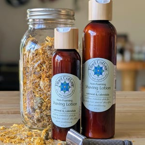 May include: A still-life image featuring two brown bottles of shaving lotion with wooden caps, a jar filled with dried flowers, and a silver razor. The lotion bottles have labels that read "Blue Cottage Arts and Apothecary."