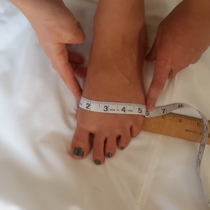 May include: A white measuring tape is used to measure the length of a person's foot. The tape measure reads 5 inches.