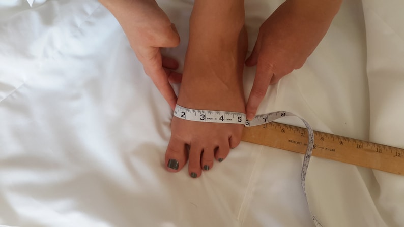 May include: A white measuring tape is used to measure the length of a person's foot. The tape measure reads 5 inches.