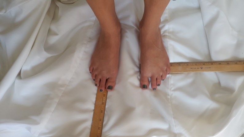 May include: A close-up of two bare feet with dark nail polish, measured with a wooden ruler. The feet are on a white fabric surface.
