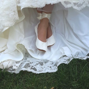 May include: White lace wedding dress with a white lace trim. The dress is pulled up to reveal a pair of white lace bridal shoes with white satin ribbons.
