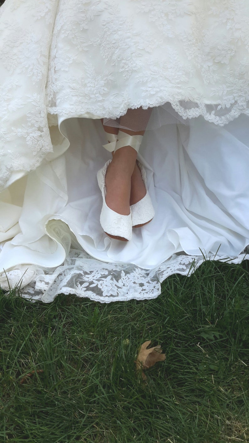 May include: White lace wedding dress with a white lace trim. The dress is pulled up to reveal a pair of white lace bridal shoes with white satin ribbons.