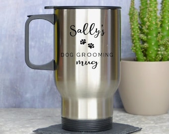 May include: Stainless steel travel mug with a black handle and lid. The mug has the text "Sally's DOG GROOMING mug" in black. Two paw prints are above the words "dog grooming". The mug is on a square black coaster.