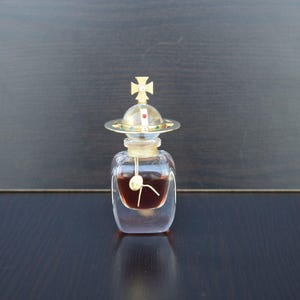 Op de afbeelding: Een heldere glazen fles met een gouden dop en een kruisontwerp. De fles bevat een donkere vloeistof en staat op een donker oppervlak.